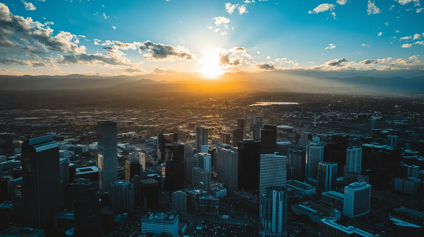 Aerial view of a Southern California city skyline with high-rise buildings at sunset, with mountains visible in the distance and sunlight streaming through clouds.